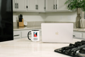 Photo of a mac laptop and a Krista Hopkins Homes branded coffee mug sitting on a kitchen counter